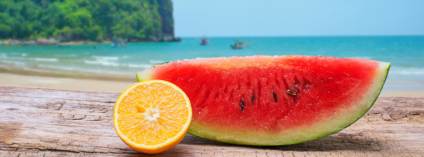 A cut watermelon with a sliced orange by the beach